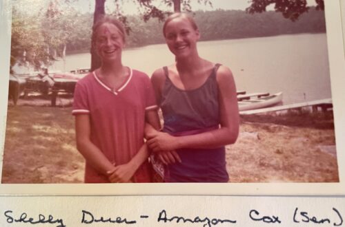 A vintage photo of two smiling campers at Camp Nakanawa, standing near Lake Aloaloa. Shelly Duer, the Amazon War Canoe cox, sports a painted “A” on her forehead—marking the beginning of a beloved camp tradition.